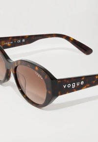 Occhiali da sole tartaruga con lenti marroni. Presentano una montatura spessa e tempie larghe, con il marchio "vogue" stampato in bianco sulle aste.