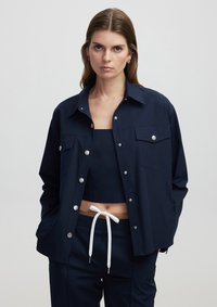 Marineblauwe jas met zilveren drukknoopjes, twee borstzakken en een relaxte pasvorm; gedragen over een bijpassende marineblauwe crop top en broek met een witte koord.
