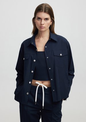 Námorně modrá bunda se stříbrnými cvoky, dvěma náprsními kapsami a volným střihem; nošena přes odpovídající námořnický crop top a kalhoty s bílým šňůrkovým pasem.