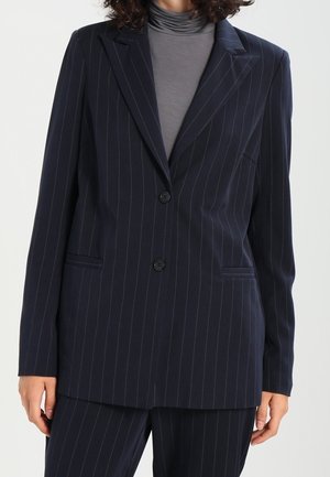 Person iført en mørk navy pinstripe blazer med to knapper over en grå rullekrave, matchende pinstripe bukser synlige nederst.