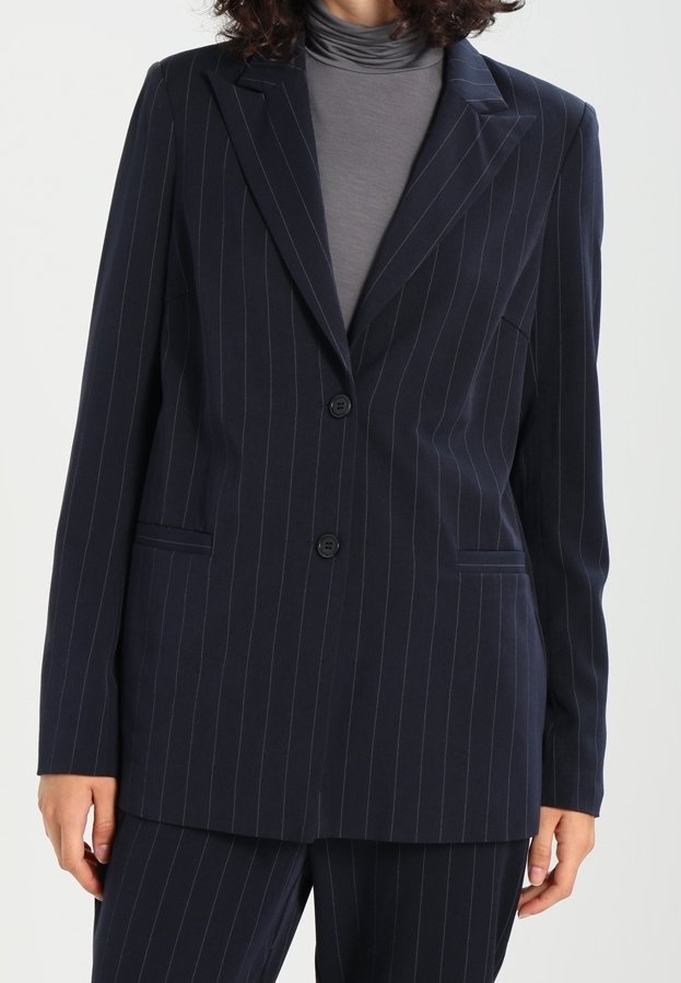 Personne portant un blazer bleu marine foncé à fines rayures avec deux boutons sur un col roulé gris, avec un pantalon assorti à fines rayures visible en bas.