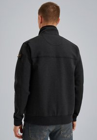 Homme portant une veste zippée gris foncé avec un col montant, debout de dos à la caméra devant un fond uni.