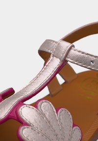 Κοντινό πλάνο σανδαλιού παιδιού με μεταλλικές ασημί λουριά και ροζ διακοσμητικό σε σχήμα λουλουδιού σε καφέ σόλα.