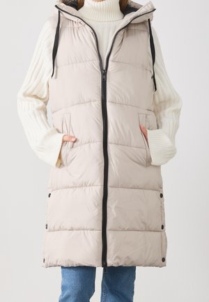 Nainen beige tikattu hupullinen toppaliivi päällään, valkoinen pooloneule ja siniset farkut, kädet liivin taskuissa.