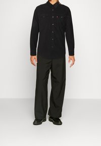 Chemise en velours côtelé noir à manches longues, boutonnée sur le devant, avec deux poches poitrine et des empiècements discrets aux épaules ; portée avec un pantalon noir à jambes larges et des bottes.