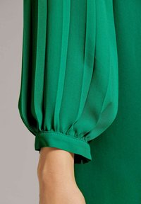 Grüne Bluse mit gefältelten langen Ärmeln. Weicher, fließender Stoff mit elastischen Bündchen. Geschmeidige Textur und lebendige Farbe.