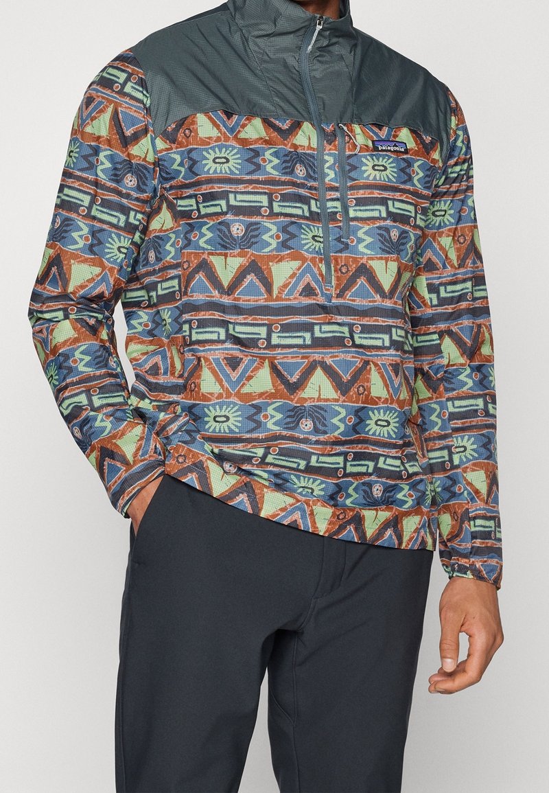 Pullover jakken har et farverigt geometrisk mønster i blå, grøn og orange, med en mørkegrå øverste del og frontlynlås.