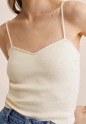 Frau trägt ein eng anliegendes cremefarbenes, geripptes Tanktop mit dünnen Trägern und hoch taillierte blaue Jeans vor neutralem Hintergrund.