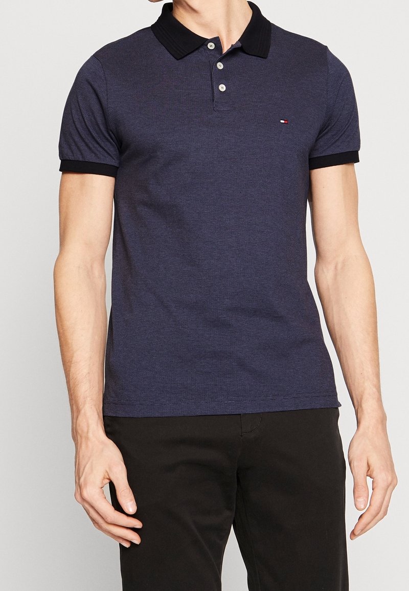 Homme portant un polo bleu marine à manches courtes avec col et poignets noirs, associé à un pantalon noir, debout devant un fond uni.