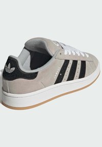 Grauer Veloursledersneaker mit schwarzen Mesh-Akzenten, weißen Schnürsenkeln, drei schwarzen Streifen und einer Gummisohle in Kaugummifarben. Verfügt über ein Adidas-Logo an der Ferse.