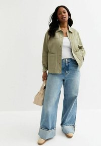 Chaqueta verde oliva con botones a presión, combinada con jeans de mezclilla de pierna ancha en un tono azul claro. Un bolso beige y zapatos en color tostado completan el atuendo.