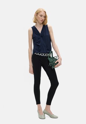 Młoda kobieta ubrana w granatowy skręcany top bez rękawów, czarne dopasowane spodnie z srebrnym paskiem, szare obcasy, trzymająca zieloną torebkę z koralików.