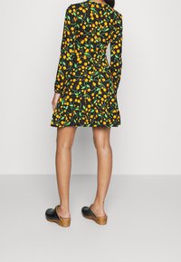 Femme portant une robe noire avec un motif de fruit orange et de feuille verte, debout, vue de dos, portant des sabots noirs à semelles en bois.