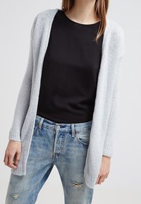 Jasnoszara dziergana kardiganka z otwartym przodem, długimi rękawami i prążkowanymi mankietami, noszona na czarną bluzkę i postrzępione niebieskie dżinsy.