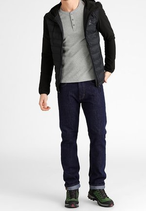 Mand iført sort hættetrøje, grå henley skjorte, mørkeblå jeans og grønne vandrestøvler står imod hvid baggrund.