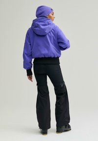 Lila kapucnis dzseki rugalmas szegéllyel, laza fekete sí nadrággal, amely állítható pántokkal rendelkezik, fekete csizmával, hátulnézetben.