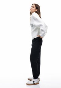 Hvid sweatshirt med grafisk tryk, sorte sweatpants og hvide sneakers med sorte accenter. Model poserer sidelæns mod en hvid baggrund.