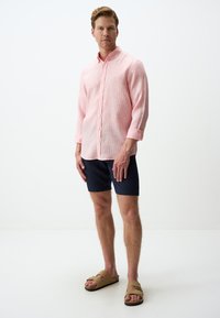 Helles rosa Hemd mit einem strukturierten Muster, kombiniert mit marineblauen Shorts und beigen Sandalen. Das Hemd hat hochgekrempelte Ärmel und einen Kragen.