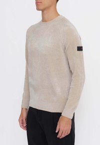 Beiger Strickpullover mit strukturierten Schulternähten und gerippten Bündchen. Auf dem linken Ärmel befindet sich ein schwarzes Patch mit Logo.