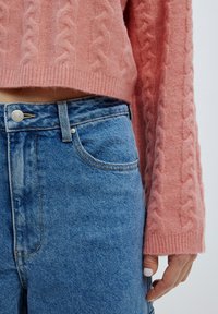 Rosa Kabelstrickpullover mit rippenförmigem Saum, kombiniert mit blauen, hoch taillierten Jeans aus Denim, die einen silbernen Knopf und sichtbare Taschennaht haben.