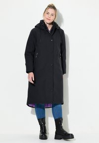 Svart vattentålig lång jacka med huva, med knappar och zippered sidofickor; fodrad med lila tyg. Bärs med blå jeans och svarta stövlar.