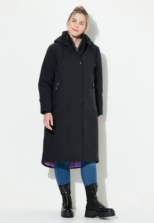 Black waterproof long coat with a hood, featuring buttons and zippered side pockets; lined with purple fabric. Worn with blue jeans and black boots.