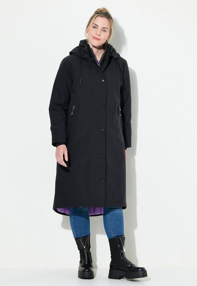 Svart vattentålig lång jacka med huva, med knappar och zippered sidofickor; fodrad med lila tyg. Bärs med blå jeans och svarta stövlar.