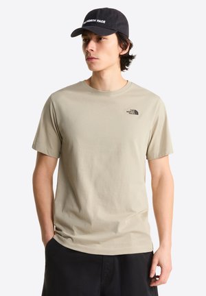 Junger Mann, der ein beiges The North Face T-Shirt und eine schwarze Kappe trägt, steht mit einer Hand in der Tasche vor einem schlichten Hintergrund.