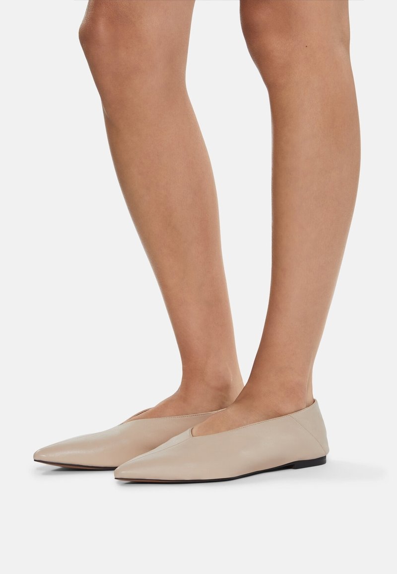 Jambes nues portant des chaussures plates beiges à bout pointu avec un petit talon, présentées sur un fond blanc.