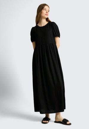 Frau steht und trägt ein langes schwarzes Kleid mit aufgebauschten kurzen Ärmeln und schwarze offene Sandalen, schaut seitlich vor einem einfarbigen Hintergrund.