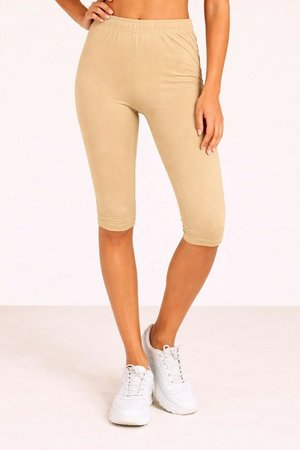 Femme portant des leggings beige ajustés au niveau des genoux et des baskets blanches, debout avec une main posée sur la hanche sur un fond uni.