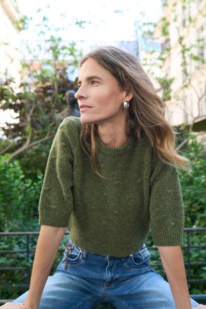 Femme aux longs cheveux portant un pull court en maille verte et un jean bleu, assise à l'extérieur, regardant sur le côté avec de la verdure et des bâtiments en arrière-plan.