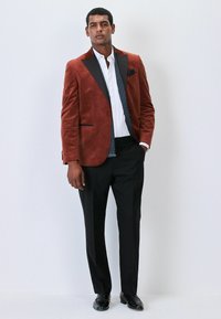 Rustfarbenes Samtblazer mit schwarzen Revers, kombiniert mit einem weißen Hemd und schwarzen Hosen. Der Blazer verfügt über Taschen und einen strukturierten Stoff.