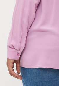 Gaiši rozā blūze ar garām, nedaudz pūkainām piedurknēm, pogu aprocēm un mīkstu tekstūru, apvienota ar ziliem džinsiem.