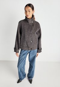 Kvindelig iført en højhalset knappet ternet jakke, bredbenede blå jeans og spidse sorte sko med metaløjer, stående på en ensfarvet baggrund.