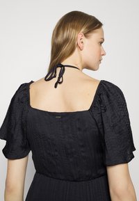 Μαύρο φόρεμα με κοντούς φουσκωτούς μανίκους και τετράγωνη λαιμόκοψη στην πλάτη. Διαθέτει υφή υφάσματος και κλείσιμο με κορδόνι στον πίσω λαιμό.