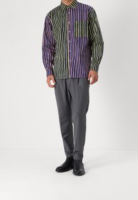 Overhemd met knoopsluiting in gestreept patroon met verticale lijnen in paars, groen en marineblauw, voorzien van een enkele borstzak. Gecombineerd met grijze broeken.