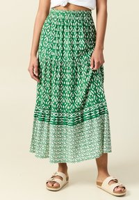 Groene tiered maxi rok met witte geometrische patronen, elastische tailleband en contrasterende patroon onderkant, gecombineerd met lichtgekleurde sandalen.
