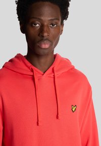 Hoodie corail orange en tissu doux, avec un capuchon à cordon de serrage et un petit logo noir et jaune sur la poitrine.