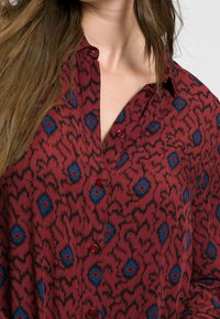 Chemise marron avec un motif floral noir et bleu, dotée d'un col et de boutons rouges. Le tissu présente une apparence douce et texturée.