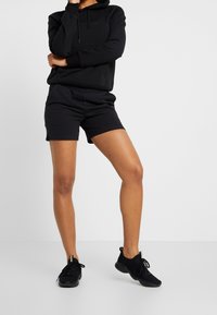 Musta huppari, jossa on kengurupussi, yhdistettynä mustiin shortsiiin. Mallilla on päällään mustat urheilukengät, joissa on tekstuuripinta ja joustavat nauhat.