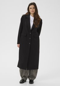 Femme debout portant un long manteau noir sur une chemise blanche, un jean ample délavé à l'acide, des chaussures noires et des boucles d'oreilles créoles dorées, devant un fond uni.