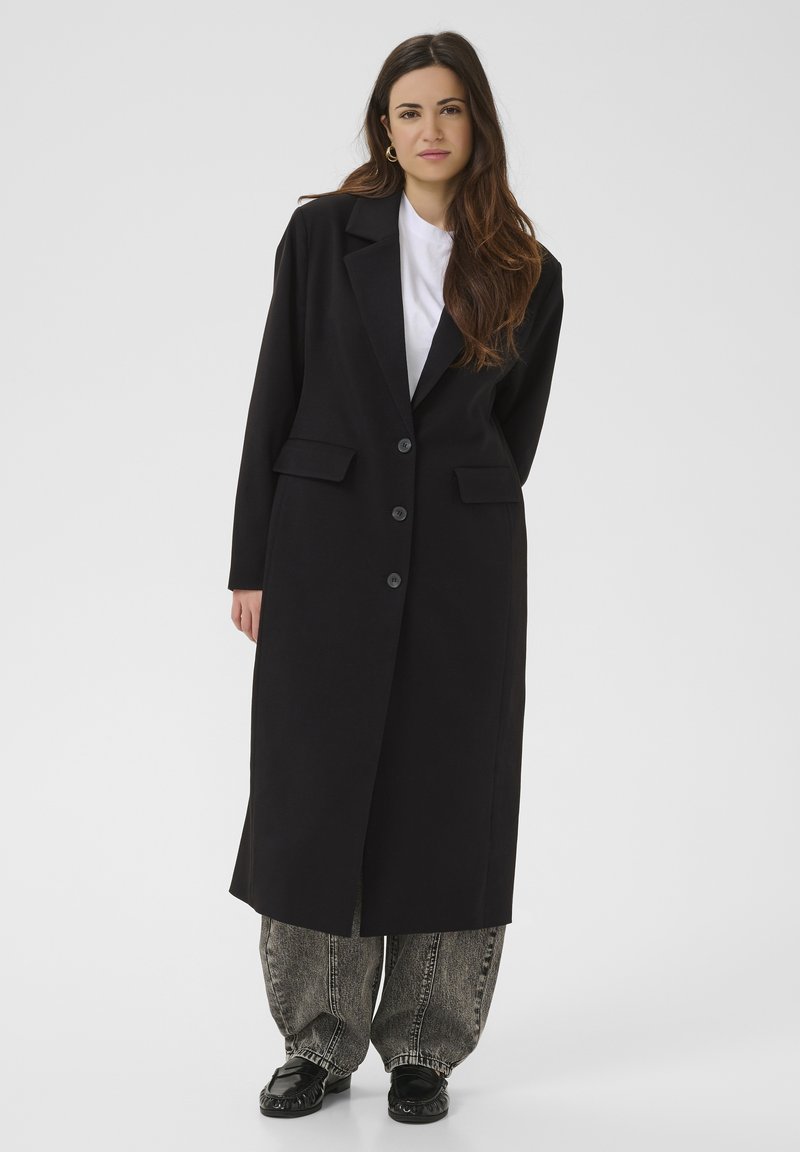 Femme debout portant un long manteau noir sur une chemise blanche, un jean ample délavé à l'acide, des chaussures noires et des boucles d'oreilles créoles dorées, devant un fond uni.