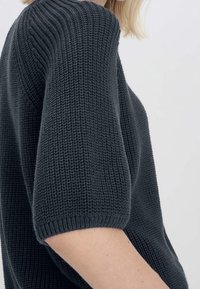 Gros plan sur une personne portant un pull épais en maille gris foncé avec des manches jusqu'aux coudes, sur un fond clair uni.