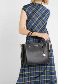 Borsa a mano in pelle nera con finitura texture, doppio manico e hardware in tonalità oro, inclusi un lucchetto e una catena decorativa.