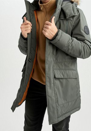 Mann trägt einen grauen Wintermantel mit Kapuze und Pelzbesatz sowie einen braunen Reißverschluss-Pullover, hält den Mantel geöffnet, um das orangefarbene Futter zu zeigen.