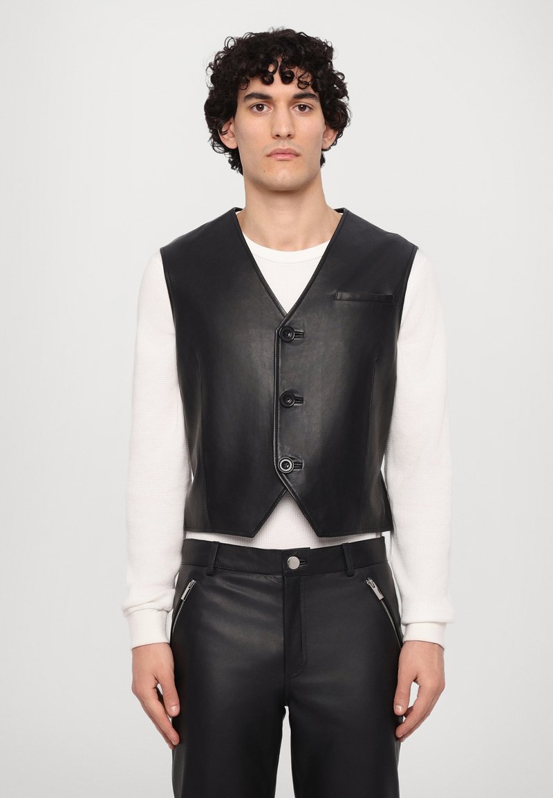 Gilet en cuir noir avec un col en V, doté d'une fermeture devant avec trois boutons et une petite poche, porté sur un haut à manches longues blanc.