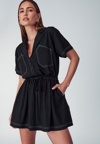 Donna che indossa un vestito nero a maniche corte con cuciture bianche, davanti con bottoni, vita con cordoncino e una mano in tasca, su uno sfondo semplice.