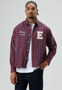 Rood en marineblauw geruit overhemd met lange mouwen, voorzien van een geborduurde "E" en de tekst "See you in hell" aan de voorkant. Witte knopen.