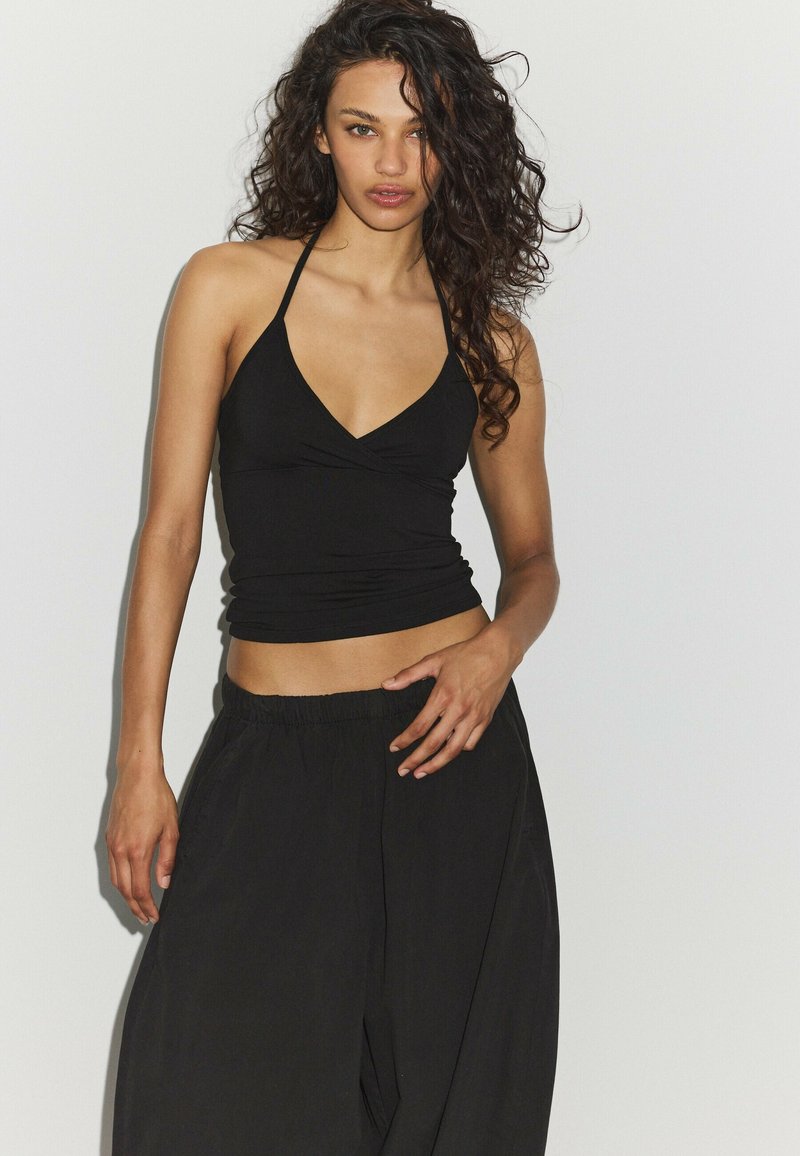 Femme aux longs cheveux bouclés portant un top court noir à col halter et un pantalon noir ample, debout devant un fond clair uni.